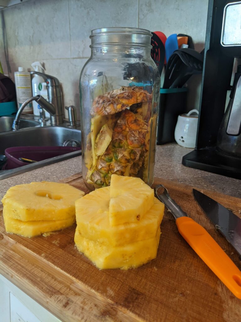 Large 2L mason jar on a cutting board. Pineapple rinds and brown liquid in the jar with a fermentation lid on top. Slices of pineapple in front of the jar on the cutting board.