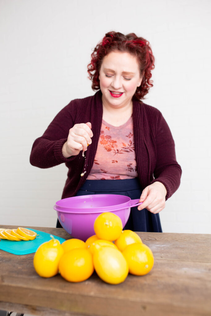 Heather O'Shea squeezing a lemon into a purple Tupperware bowl. A pile of lemons sit in front of the bowl, and a turquoise cutting board with a sliced lemon on it sits to the left of the bowl.