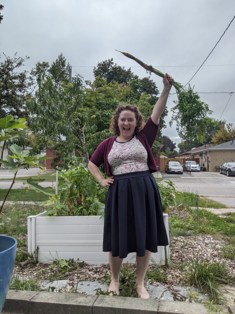 Heather O'Shea, standing in front of a raised, white, garden bed, holding a large carrot above her head. 