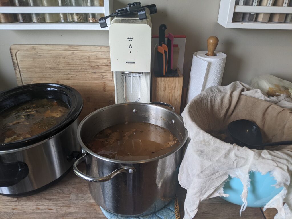 Slow cooker, a stock pot, and a large Tupperware bowl with a strainer and tea towel on top.