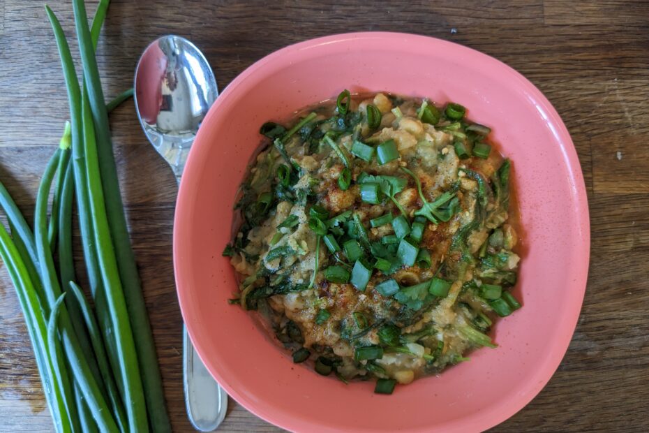 A salmon pink coloured bowl full of cumin stewed chickpeas, topped with cilantro and green onions on a wood countertop, next to a spoon and a bunch of green onions.