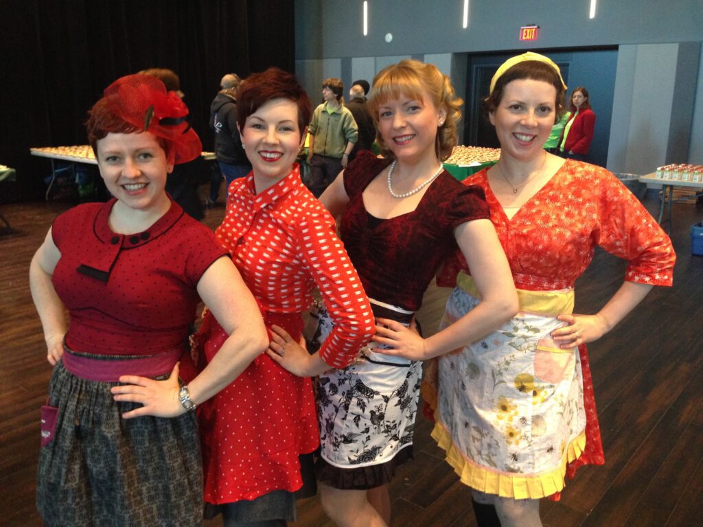 Heather O'Shea and three friends, all dressed in red vintage style dresses with vintage style aprons, standing together with one hand on their hips.