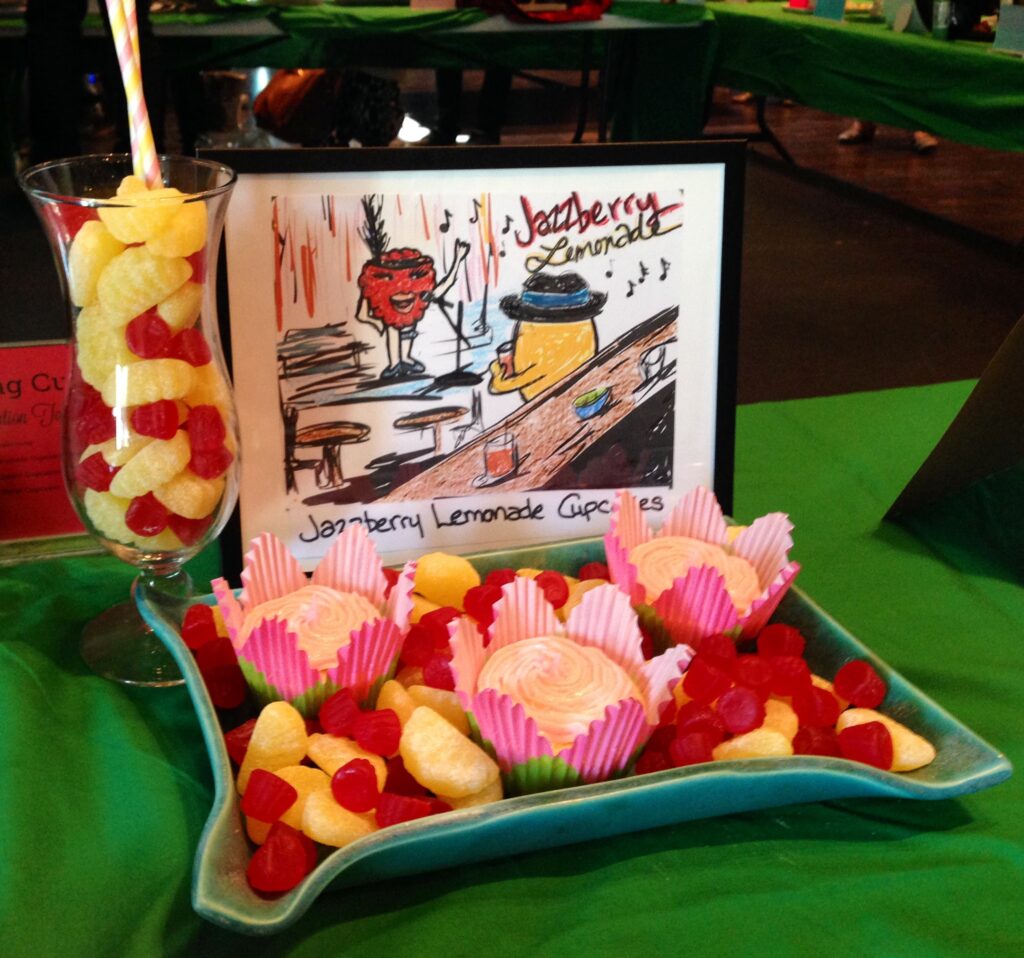 Display of cupcake entry entitles "Jazzberry Lemonade Cupcakes" with a plate of three cupcakes, and some candy rasperries & lemon slices in a cocktail glass.