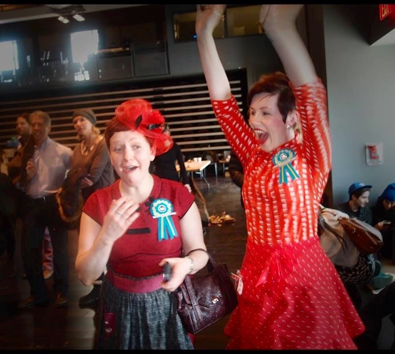 Heather and Friend. Heather's shocked face and friend jumping with arms up in excitment at the announcement that Heather's cupcakes won best in show.