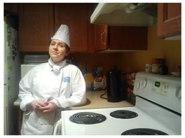 Heather O'Shea leaning on a kitchen counter, near a stovetop. She is wearing a white chef's uniform and hat.