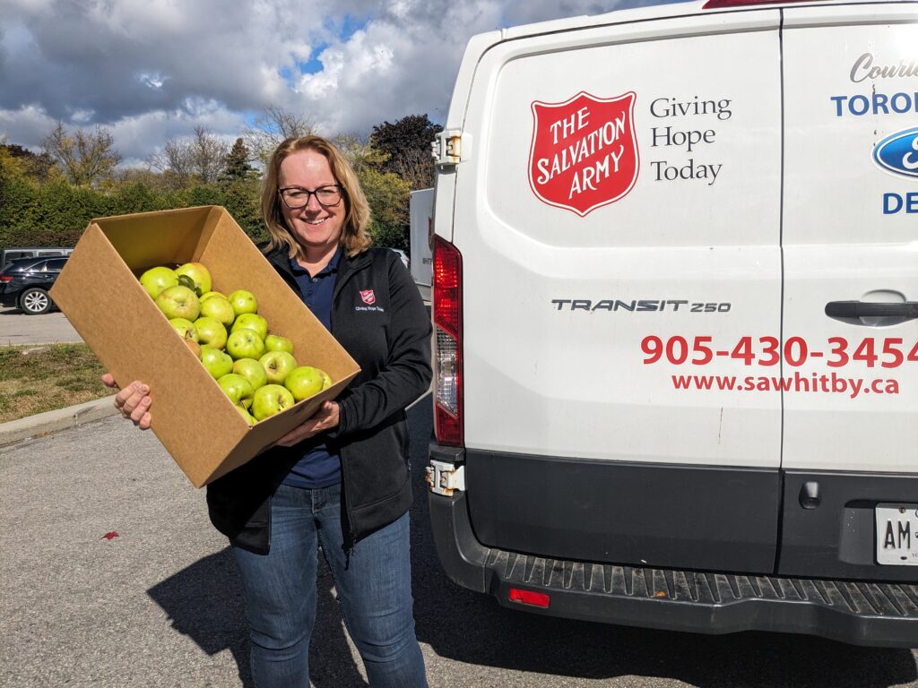 A woman with glasses and blonde hair, wearing a black Salvation Army jacket, smiles while holding a large cardboard box filled with freshly harvested green apples. She stands in front of a white Salvation Army van with red and blue lettering, displaying the organization's logo and contact information. The background features a partly cloudy sky, trees, and parked cars.