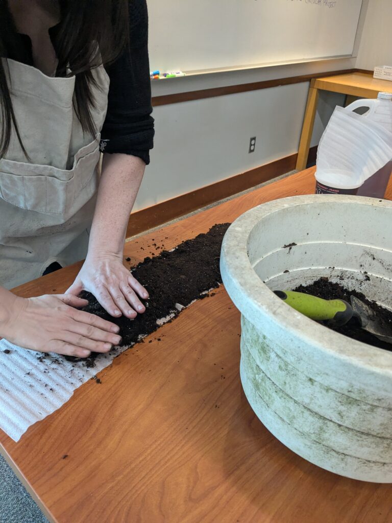A person with light skin, wearing a black top and a beige apron pressing dark, damp soil onto a wide strip of corrugated packing wrap.