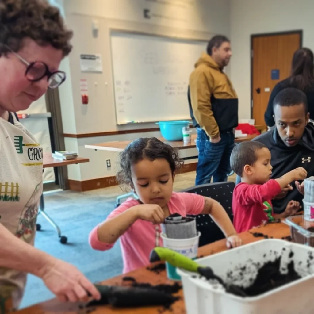 Three adults and two children standing at a table, making seed snails, taking soil from a bin and placing it onto strips of packaging material. 