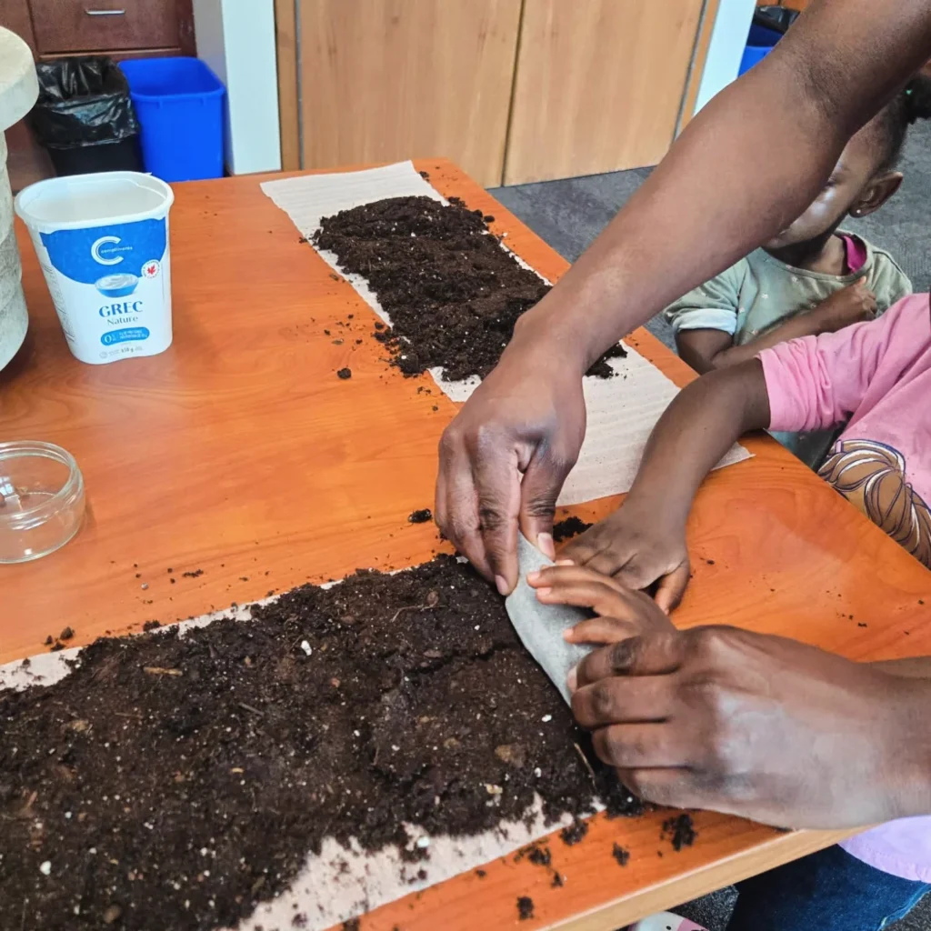 Two people with brown skin, one adult and one child, working together to begin to roll the strip of packing material covered with soil into a roll. 