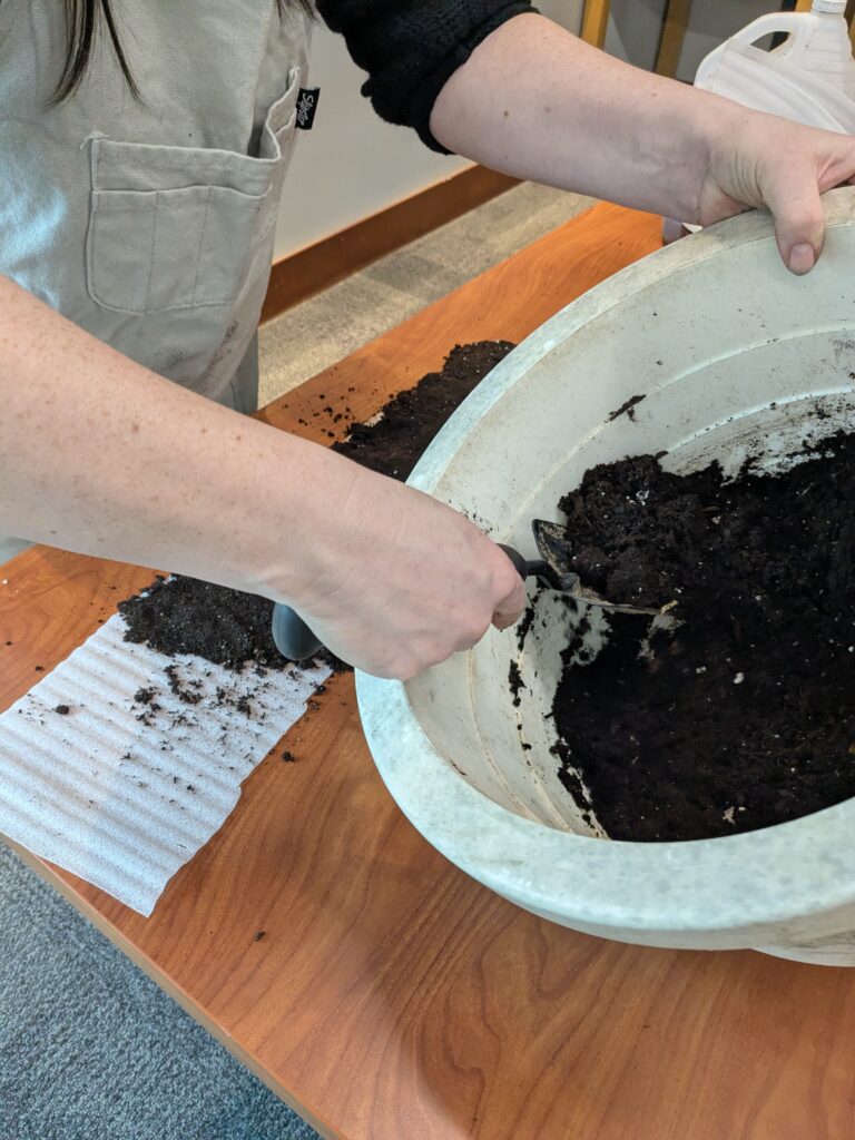 Damp, dark soil being mixed by a person with light skin using a garden trowel in a large, white bucket.