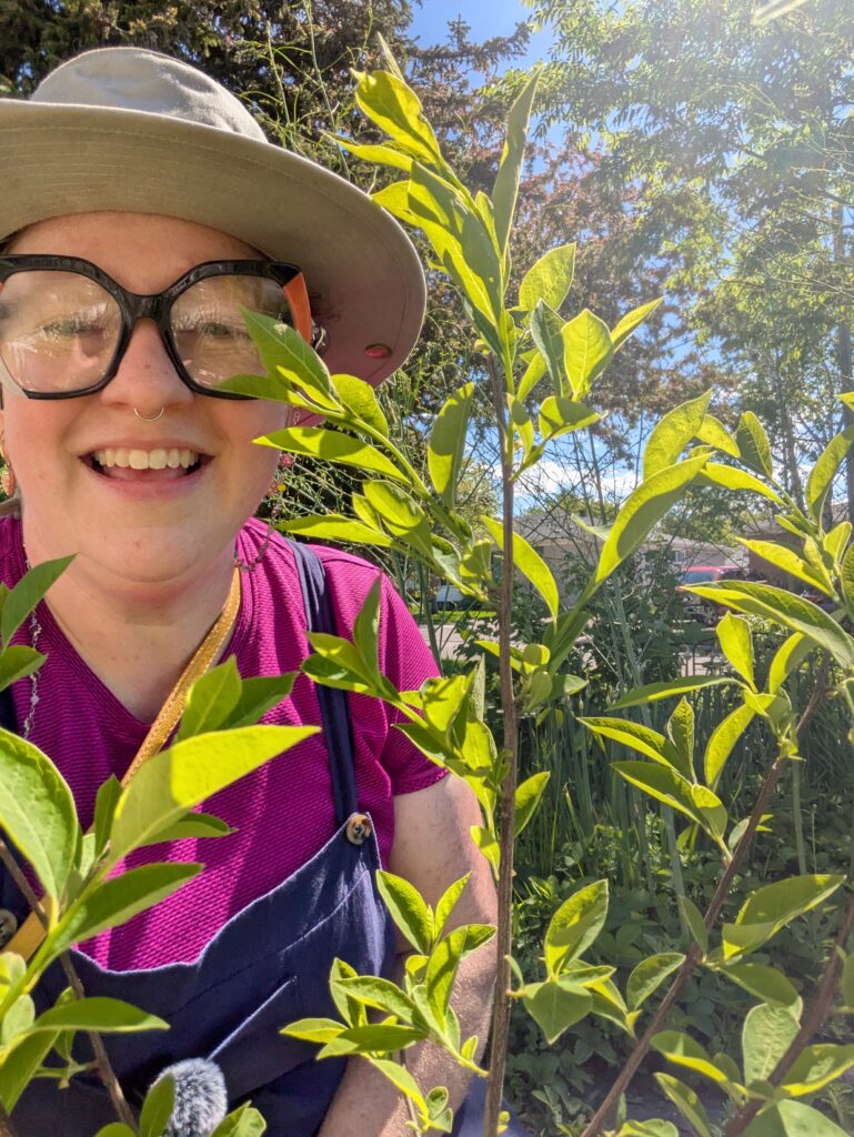 Heather, wearing a beige hat, glasses, and a pink shirt, sun shining, several branches with leaves on them in the foreground. 